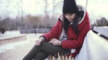 Kışın giydiği elbiselerle şehir parkında bankta oturan genç bir kadın satranç oynar. Siyah şapkalı kadın, satrançta tahta sıralarda, bulutlu havada..