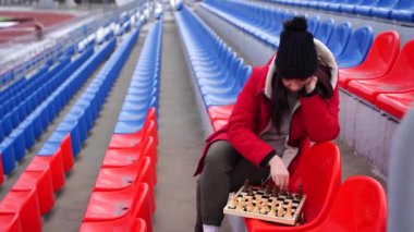 Kışlık elbiseli genç kadın satranç oynuyor, stadyum tribünlerinde tek başına oturuyor. Siyah şapkalı kadın, bulutlu havada spor stadyumunda satranç oynuyor..