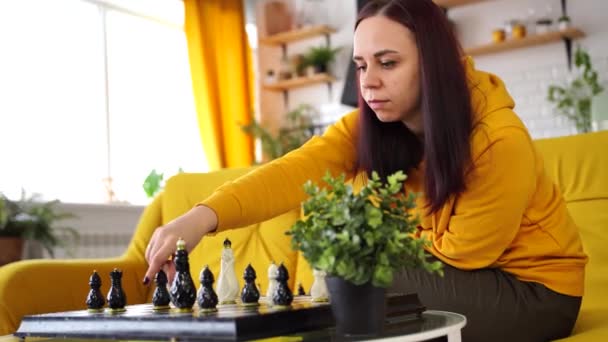 Νεαρή γυναίκα κάθεται στον κίτρινο καναπέ και παίζει σκάκι στο δωμάτιο. Γυναίκα παίζει στο λογικό επιτραπέζιο παιχνίδι με τον εαυτό της. — Αρχείο Βίντεο
