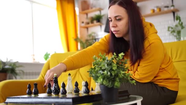 Mladá žena sedící na žluté pohovce a hrající šachy v místnosti. Žena hraje v logické deskové hře sama se sebou. — Stock video