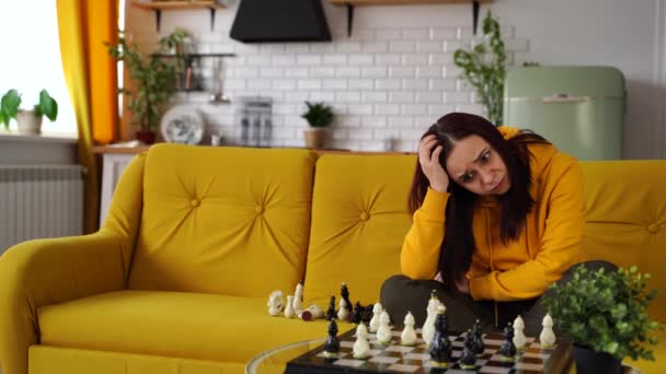 Молода засмучена жінка грає в шахи, сидячи на дивані. Занепокоєна жінка грає в логічну настільну гру з собою в кімнаті . — стокове відео