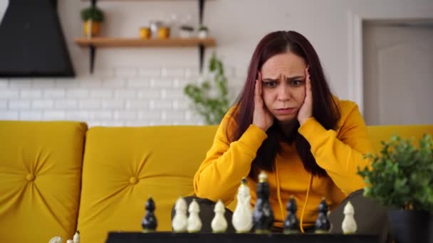 Joven mujer molesta jugando ajedrez sentado en el sofá. Mujer angustiada juega en el juego de mesa lógica consigo misma en la habitación. — Vídeo de stock