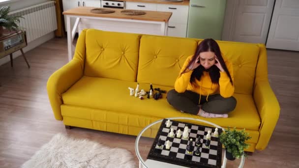 Jonge overstuur vrouw die schaak speelt zittend op de bank. Verstoorde vrouw speelt in logisch bordspel met zichzelf in de kamer. — Stockvideo