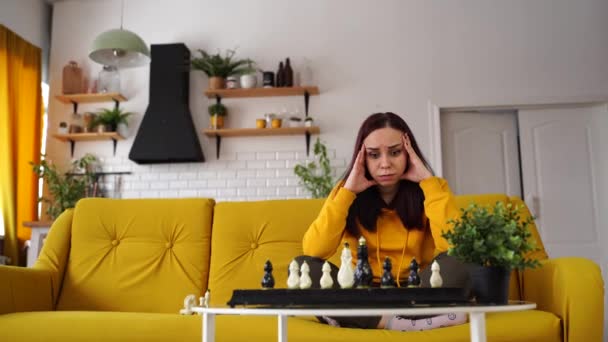 Молодая расстроенная женщина играет в шахматы, сидя на диване. Несчастная женщина играет в логическую настольную игру с самой собой в комнате. — стоковое видео