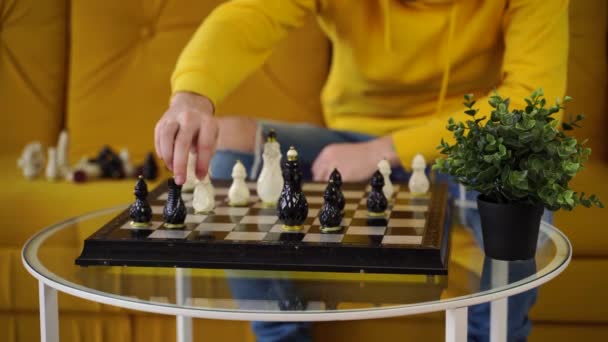 Молодий чоловік сидить на жовтому дивані і грає в шахи в кімнаті. Чоловік грає в логічну настільну гру з собою . — стокове відео