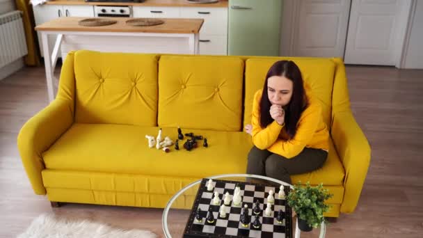 Mujer joven sentada en un sofá amarillo y jugando al ajedrez en la habitación. Hembra jugando en el juego de mesa lógica con ella misma. — Vídeos de Stock