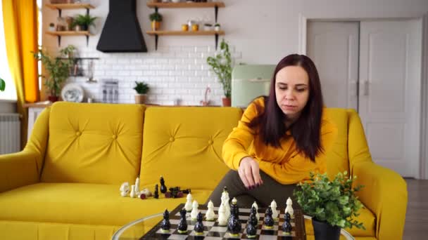 Jonge vrouw zit op een gele bank en schaakt in de kamer. Vrouwelijk spelen in logisch bordspel met zichzelf. — Stockvideo