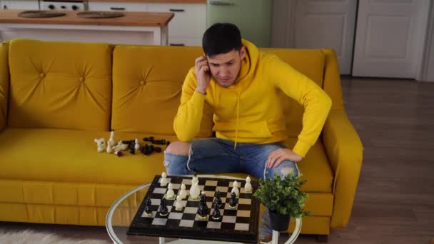 Un tânăr stând pe o canapea galbenă şi jucând şah în cameră. Bărbat care joacă în joc de bord logic cu el însuși . — Videoclip de stoc