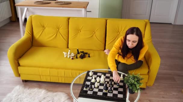 Mladá žena sedící na žluté pohovce a hrající šachy v místnosti. Žena hraje v logické deskové hře sama se sebou. — Stock video