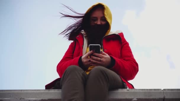 携帯電話を持つ若い女性がコンクリートのパレットに座っている 黄色のパーカーと赤いジャケットでブルネット曇りの天気でスマートフォンを閲覧 — ストック動画