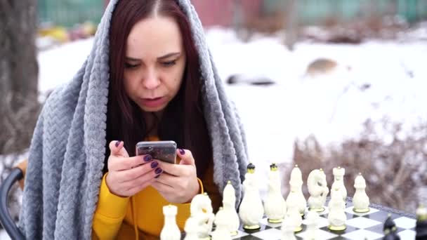 年轻女子在院子里下棋和浏览智能手机 穿着灰色格子呢衣服 手持手机坐在街上玩棋盘游戏的女性 — 图库视频影像