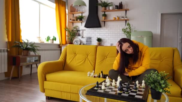 Ung upprörd kvinna spelar schack sittandes på soffan. Besvärad kvinna spelar i logiskt brädspel med sig själv i rummet. — Stockvideo