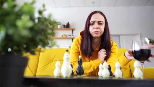 Mujer joven jugando al ajedrez y bebiendo vino tinto en la habitación. Mujer adulta con alcohol juega en un juego de mesa lógico consigo misma, sentada en el sofá. — Vídeos de Stock