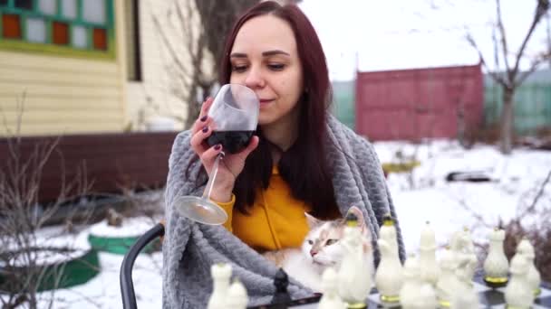 Mladá žena hraje šachy a pije červené víno na dvorku. Žena zabalená v šedé kostkované s alkoholem sedí na ulici hrát v deskové hry v zimní sezóně. — Stock video