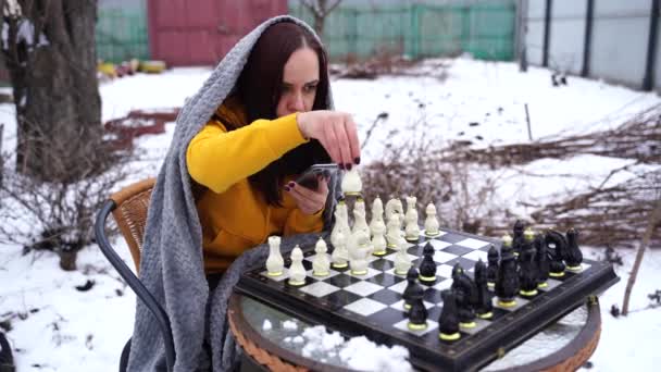 年轻女子在院子里下棋和浏览智能手机 穿着灰色格子呢衣服 手持手机坐在街上玩棋盘游戏的女性 — 图库视频影像