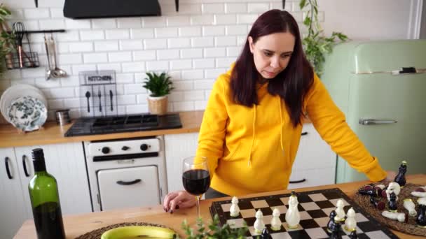 思慮深い若い女性はチェスをし 赤ワインを飲む 論理ボードゲームでアルコールプレイを持つ女性 キッチンに立って — ストック動画