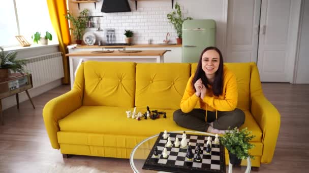 La jeune femme sourit et regarde la caméra, assise sur le canapé. Jolie femelle joue dans le jeu de société logique dans la chambre. — Video