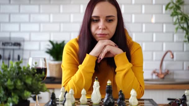 Продумана молода жінка грає в шахи, підстрибуючи підборіддя в її руках. Жінка грає в логічну настільну гру з собою на кухні . — стокове відео