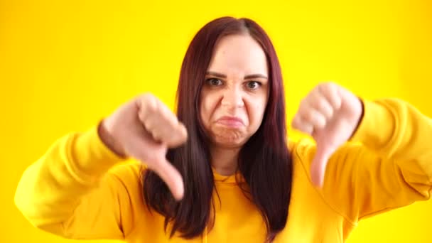 一个年轻女子面带微笑的画像，她的大拇指垂在黄色的背景上。一个穿着黄色帽衫的不快乐的黑头发的特写镜头显示了一种不赞成的姿态。概念：设置不喜欢 — 图库视频影像
