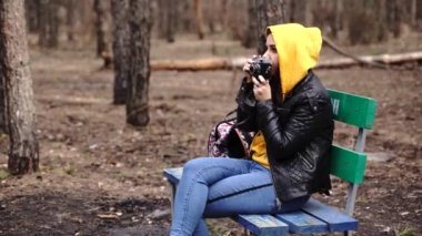 Günlük kıyafetlerle bankta oturan ve ormandaki eski fotoğraf makinesinde fotoğraf çeken genç bir kadın. Kadın dinleniyor ve bahar başında eski fotoğraf makinesiyle fotoğraf çekiyor.