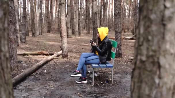 身着休闲装的年轻女子坐在长椅上 用森林里的旧相机拍照 早春女性休息及使用旧相机拍照 — 图库视频影像