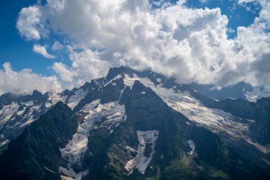 Bulutlu gökyüzüne karşı karlı dağ. Beyaz bulutların altından mavi gökyüzünde yüzen dağların üzerinden doğada güneşli bir günde karla kaplı..