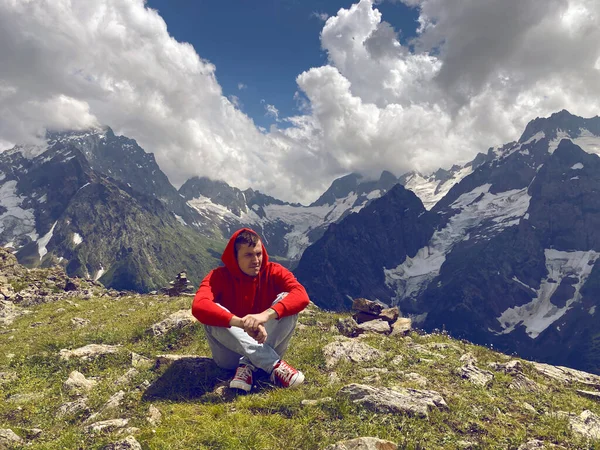 Young man sitting on mountain peak in cloudy weather. Adult guy rests, enjoying amazing mountain landscape in summertime