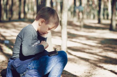 Parkta akıllı telefon kullanan odaklanmış bir çocuk. Ciddi bir şekilde konsantre olmuş, gündelik giysiler içinde okul çocuğu, sonbahar gününü parkta geçirirken bankta oturuyor ve cep telefonuyla oyun oynuyor..