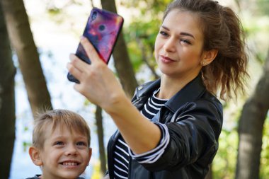 Oğluyla selfie çeken genç bir anne. Uzun saçlı ve çocuklu gülümseyen genç bir kadın parkta akıllı telefonuyla selfie çekiyor..