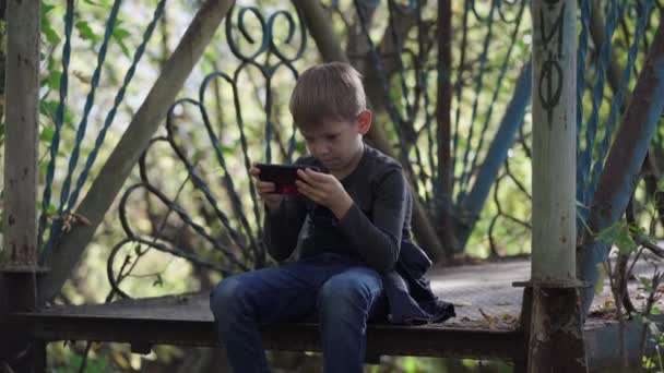 Niño enfocado usando smartphone en el parque. Niño de edad escolar concentrado serio en ropa casual sentado en el banco y jugando en el teléfono móvil mientras pasa el día de otoño en el parque — Vídeo de stock