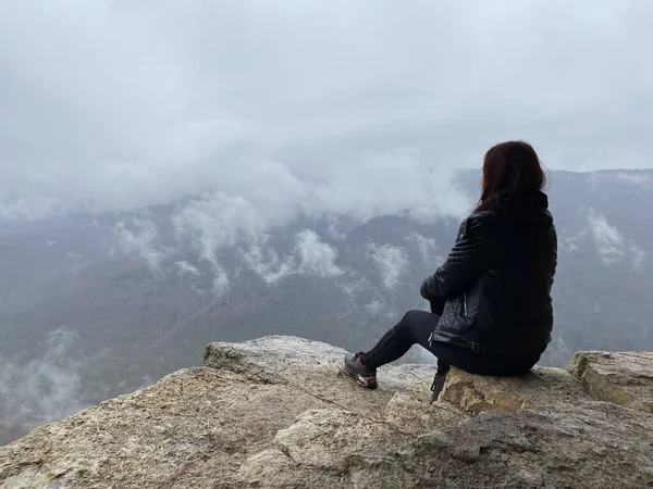 Young woman sitting on edge of cliff in foggy and cloudy weather. Female tourist enjoys spectacular view of nature, sitting on high rock