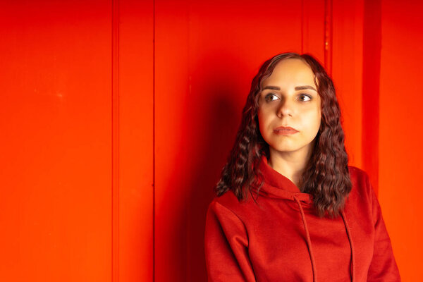 Young woman in hoodie on red background. Curly brunette poses near red wall