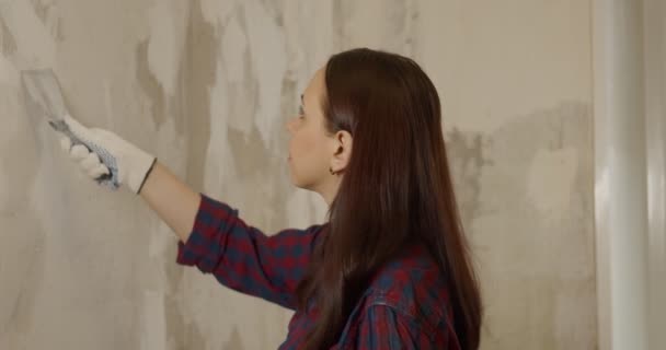 El primer plano de la hembra en guantes con espátula aplana la pared en la habitación. Mujer joven lijando la pared y preparándose para la pintura en el apartamento. Concepto de trabajos de reparación. — Vídeo de stock