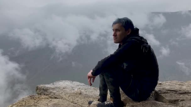 在多雾多云的天气里 一个留着蓝色头发的年轻人坐在悬崖边 男性游客坐在高高的岩石上 欣赏壮观的自然景观 — 图库视频影像