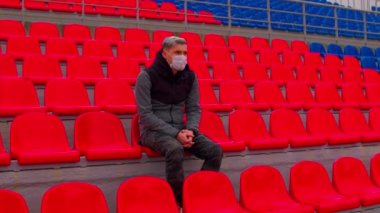 Tıbbi maskeli genç adam stadyum tribünlerinde tek başına oturuyor. Üzgün yetişkin erkek kendini spor stadyumundaki hastalıklardan koruyor. Kamu etkinlikleri ve yarışmalara kısıtlamalar.