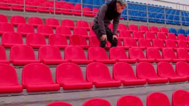 Tıbbi maskeli genç adam stadyum tribünlerinde tek başına oturuyor. Üzgün yetişkin erkek kendini spor stadyumundaki hastalıklardan koruyor. Kamu etkinlikleri ve yarışmalara kısıtlamalar.