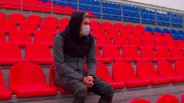 Tıbbi maskeli genç adam stadyum tribünlerinde tek başına oturuyor. Üzgün yetişkin erkek kendini spor stadyumundaki hastalıklardan koruyor. Kamu etkinlikleri ve yarışmalara kısıtlamalar.