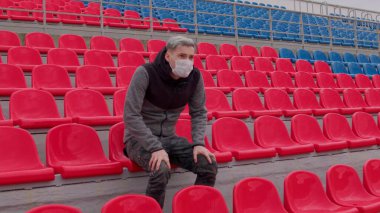 Tıbbi maskeli genç adam stadyum tribünlerinde tek başına oturuyor. Üzgün yetişkin erkek kendini spor stadyumundaki hastalıklardan koruyor. Kamu etkinlikleri ve yarışmalara kısıtlamalar