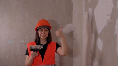 Tulum giymiş, gülümseyen ve baş parmağını kaldıran genç bir kadın. İnşaat alanında spatulası ve macunu olan kadın inşaat işçisi. Tamir işleri ve konut koşullarının iyileştirilmesi.