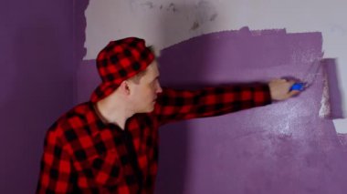 Duvarı mor renkte silindirle boyayan genç adama yaklaş. Duvara boya süren mutlu erkek..