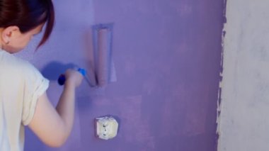 Close up of unrecognizable person painting wall in purple colour with roller. Unknown woman applying paint on wall. Concept of repair work and improvement of housing conditions