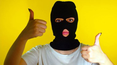 Siyah kar maskeli, sarı arka planda baş parmağını gösteren tanınmamış bir kadının yakın çekimi. Kimliği belirsiz bir kadın kameraya bakıyor ve onaylama hareketi yapıyor.