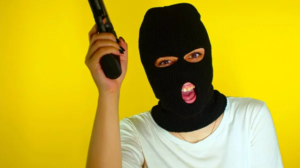 Unrecognizable woman in black balaclava with gun on yellow background. Dangerous female in mask takes aim at camera, shoots and laughs. Slow motion.