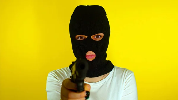 Unrecognizable woman in black balaclava with gun on yellow background. Dangerous female in mask takes aim at camera and shoots. Slow motion.