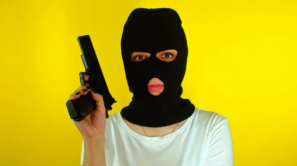 Unrecognizable woman in black balaclava with gun on yellow background. Dangerous criminal in mask with pistol looking at camera