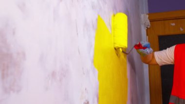 Koruyucu eldiven giymiş, sarı renkli duvarları silindirle boyayan bir kadının elini kapat. Duvara boya sürerken tanınmayan biri. Tamir işleri ve konut koşullarının iyileştirilmesi.