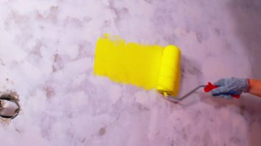 Koruyucu eldiven giymiş, sarı renkli duvarları silindirle boyayan bir kadının elini kapat. Duvara boya sürerken tanınmayan biri. Tamir işleri ve konut koşullarının iyileştirilmesi.