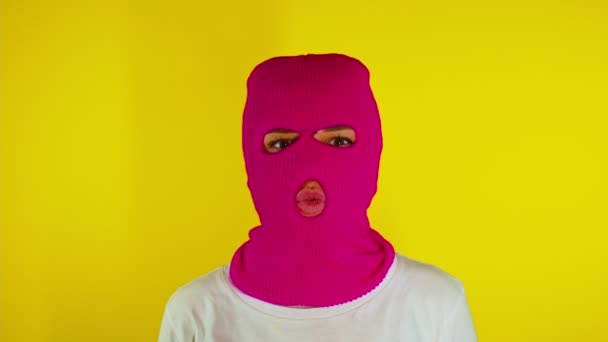 黄色の背景にキスを吹いて送信ピンクのバラクラバの認識できない女性 知らない女性でマスク見ますカメラと作りますアヒル顔とともにキス — ストック動画