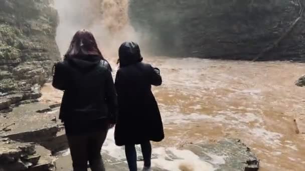 Visão traseira de duas mulheres olhando para a poderosa cachoeira lamacenta. Turistas em pé sobre rochas em terreno montanhoso e desfrutando de uma bela vista da catarata. — Vídeo de Stock