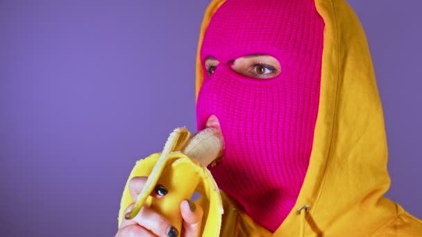 穿着粉色巴拉克拉瓦和黄色头巾的年轻女子的近照吃着紫色背景的香蕉 蒙面的不明女性咬着美味的水果 回头看 — 图库视频影像